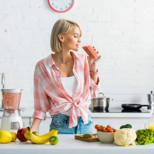 attractive blonde woman drinking tasty smoothie near ingredients in kitchen