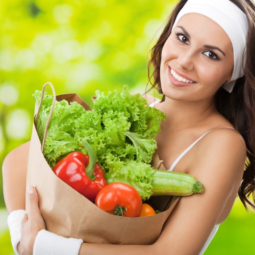 Woman in fitness wear with vegetarian food Portrait of happy smiling young beautiful woman in fitness wear holding grocery shopping bag with healthy vegetarian food, outdoors